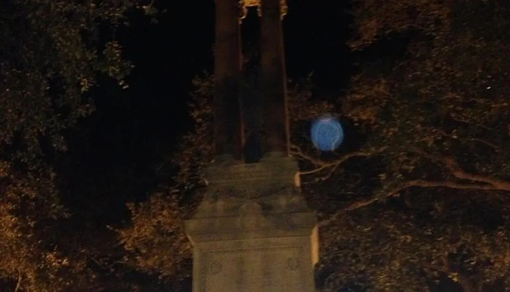 The image shows a nighttime scene with a monument or statue flanked by trees and an unusual blue orb-like feature that could be a result of the cameras lens flare or light reflection