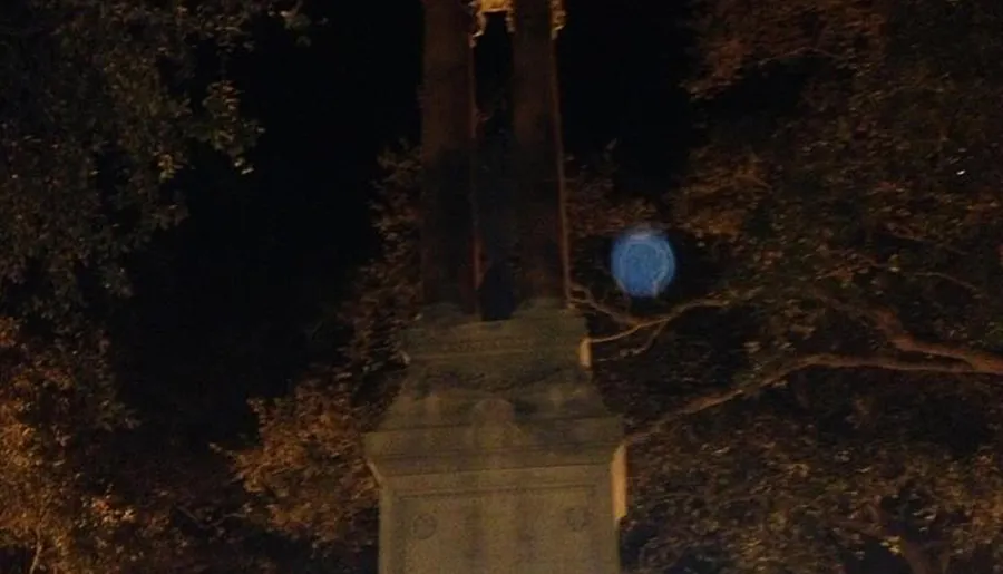 The image shows a nighttime scene with a monument or statue flanked by trees and an unusual blue orb-like feature that could be a result of the camera's lens flare or light reflection.