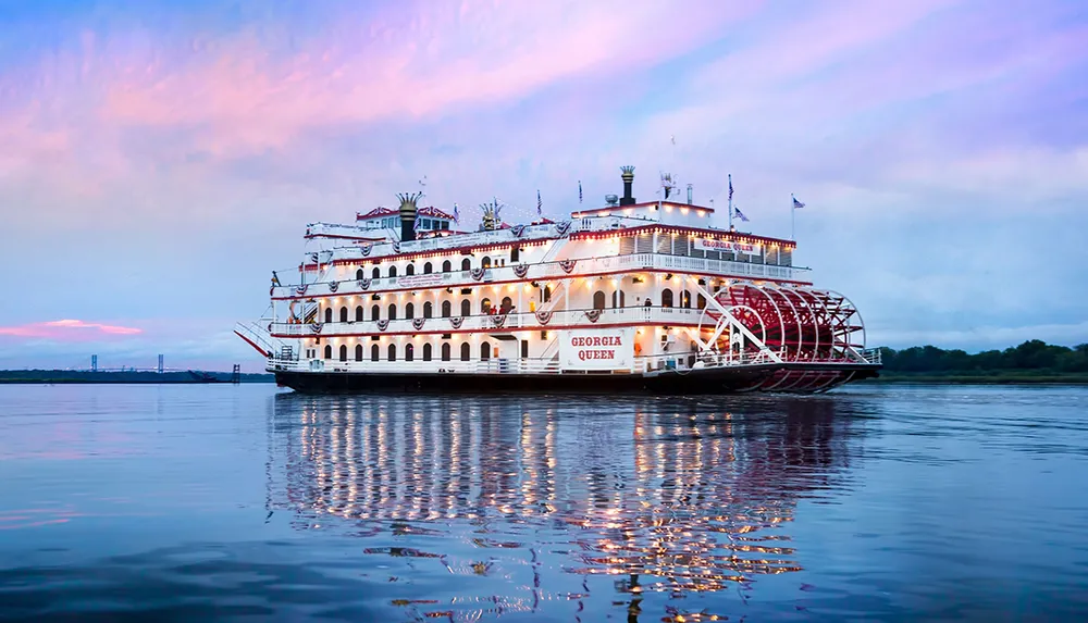 A large paddlewheel riverboat named Georgia Queen is floating on calm waters with a pastel-colored sky in the background
