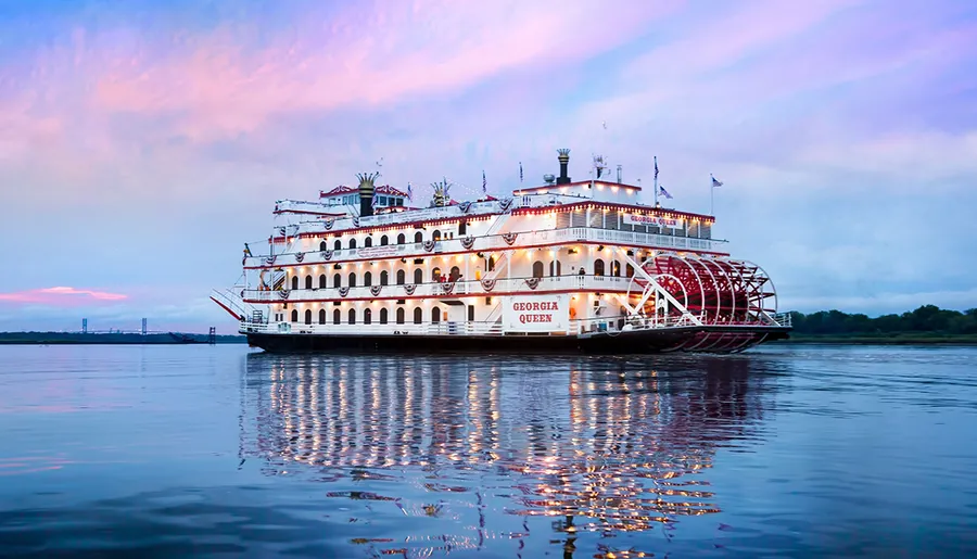 A large paddlewheel riverboat named Georgia Queen is floating on calm waters with a pastel-colored sky in the background.