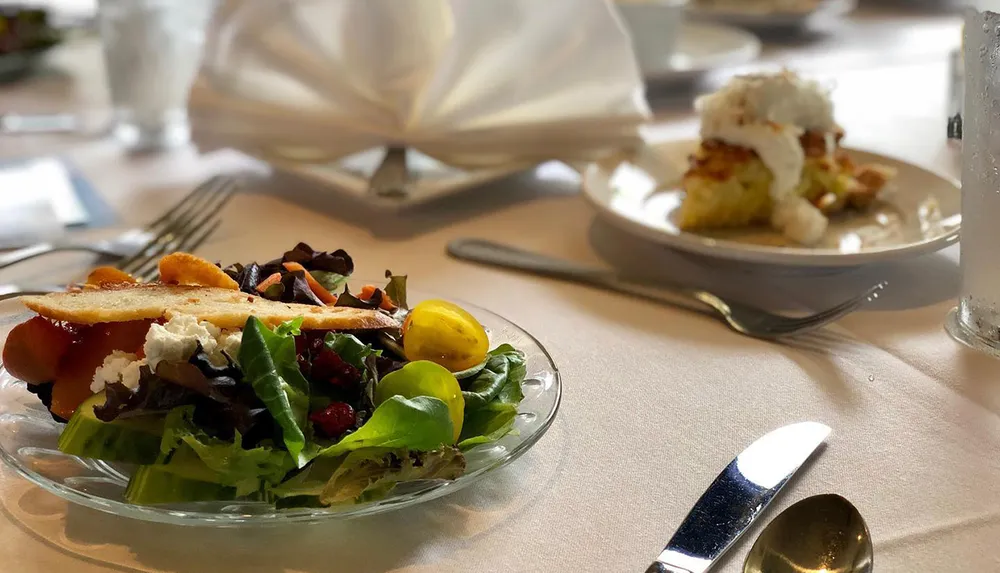 The image shows an elegantly set dining table with a leafy salad and a dessert in soft focus on the background