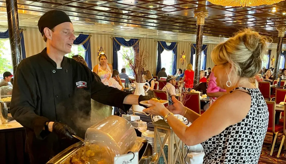 A chef is serving food to a woman at a buffet in an elegantly decorated dining room filled with guests