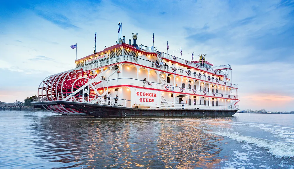 The image shows the Georgia Queen a large paddlewheel riverboat beautifully illuminated and adorned with festive lighting gliding along the water at twilight