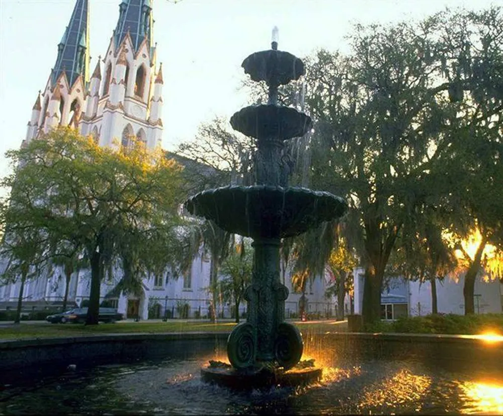 The image depicts a multi-tiered fountain in the foreground with water cascading down its levels set against the backdrop of a white Gothic-style church partially obscured by trees captured at a moment when the sun casts a warm glow through the branches