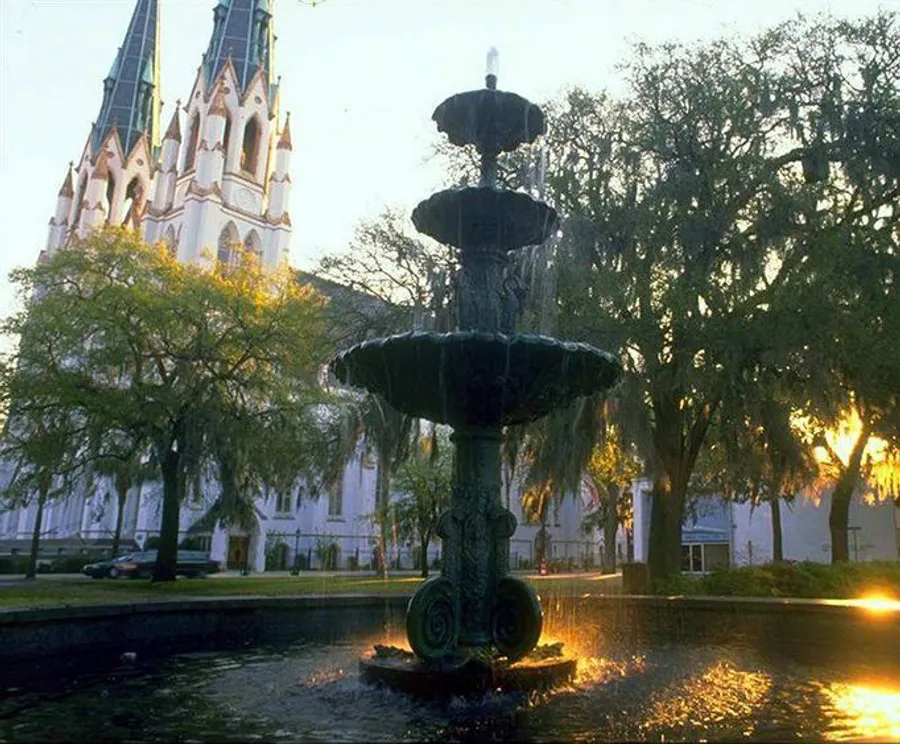 The image depicts a multi-tiered fountain in the foreground with water cascading down its levels, set against the backdrop of a white Gothic-style church partially obscured by trees, captured at a moment when the sun casts a warm glow through the branches.