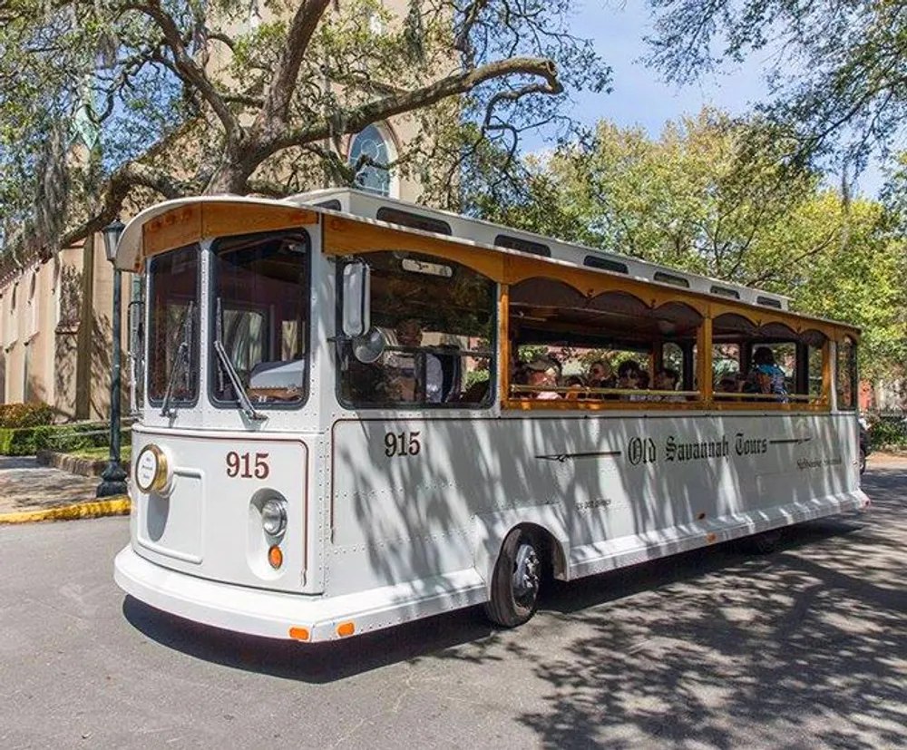 A trolley-style tour bus labeled Old Savannah Tours is filled with passengers and is driving along a tree-lined street