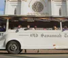 A group of people are riding a white trolley labeled Old Savannah Tours near a historic building with a sign for the Pirates House