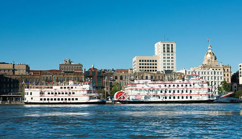 The image shows two large paddle wheel riverboats docked at a riverbank with a backdrop of a city skyline under a clear blue sky