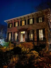A stately two-story house with warm interior lighting is photographed at night, showcasing its classic architecture and lush garden under a twilight sky.