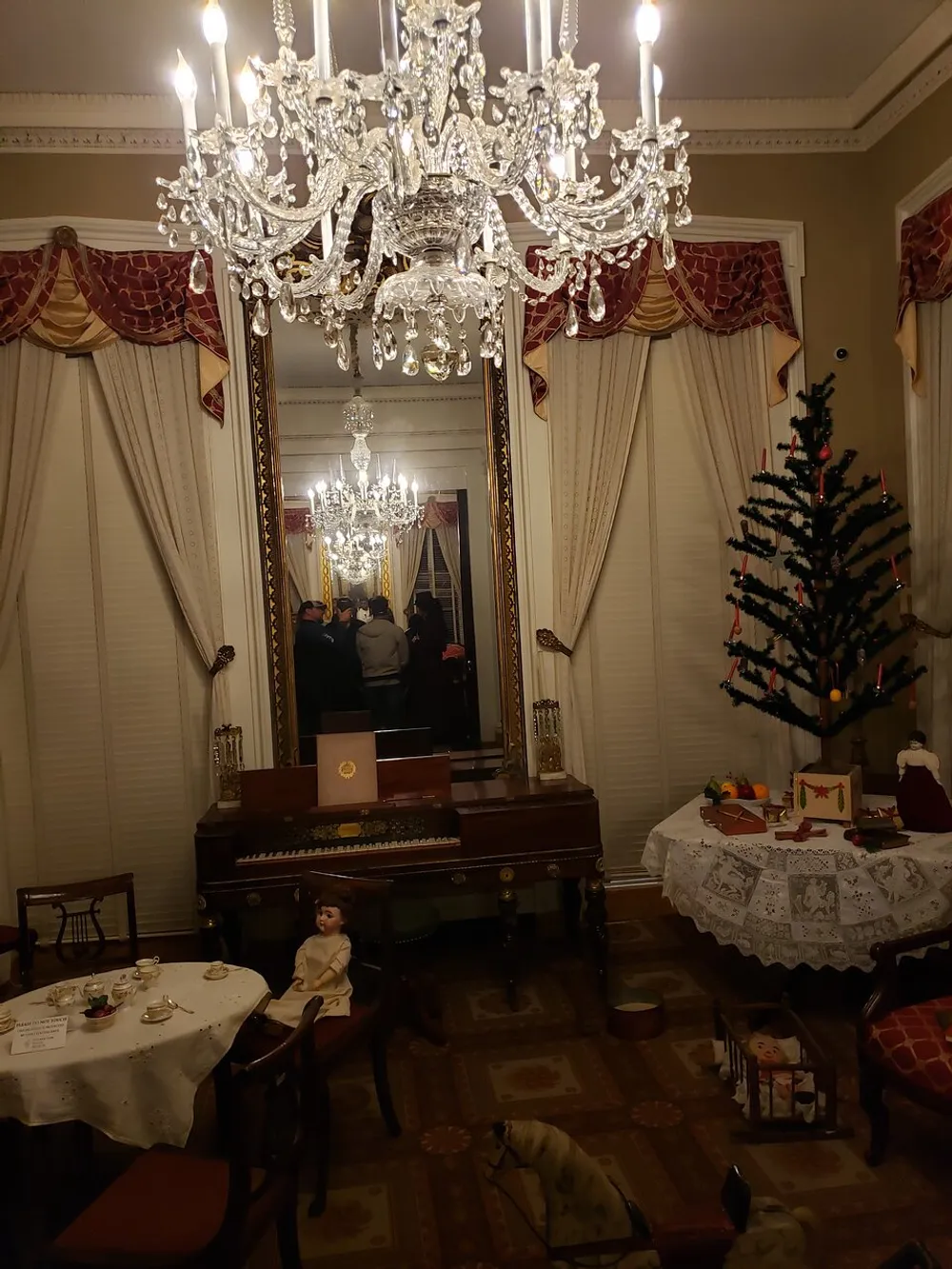 The image shows an elegantly decorated room with a large crystal chandelier ornate curtains a piano a small Christmas tree and vintage toys evoking an atmosphere of historical luxury