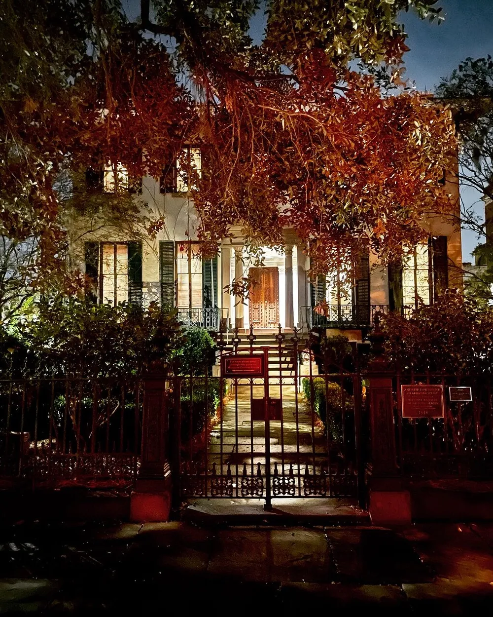 A warmly lit stately house peeks through dense foliage behind an ornate iron gate in the evening