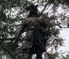 The image shows a statue of a historical figure in traditional attire including a tricorne hat and a sword set against a backdrop of leafy trees