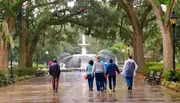 People with umbrellas and raincoats walk towards a fountain on a wet path lined with trees and street lamps, on what appears to be a rainy day.