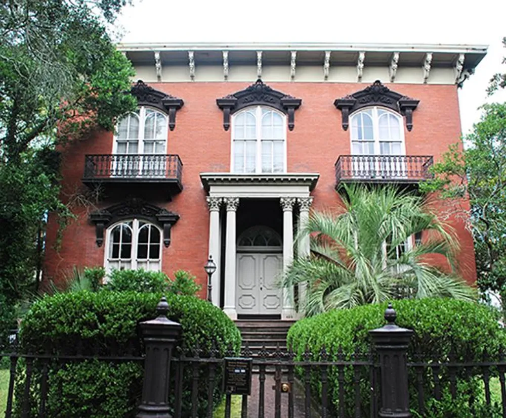 The image shows a two-story red brick historic house with black wrought iron railings arched windows ornamental cornices and a front porch surrounded by lush greenery and a metal fence