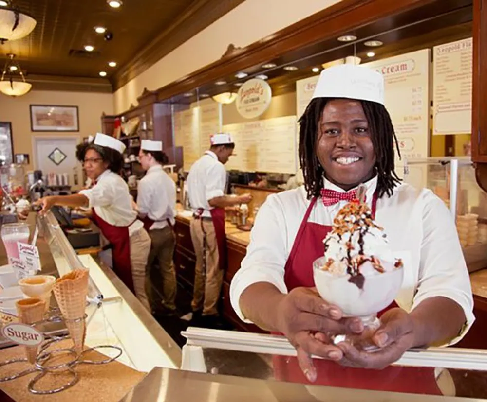 A smiling individual in a chefs hat and red apron presents a large and elaborately topped ice cream sundae at an ice cream parlor counter with other staff members working in the background