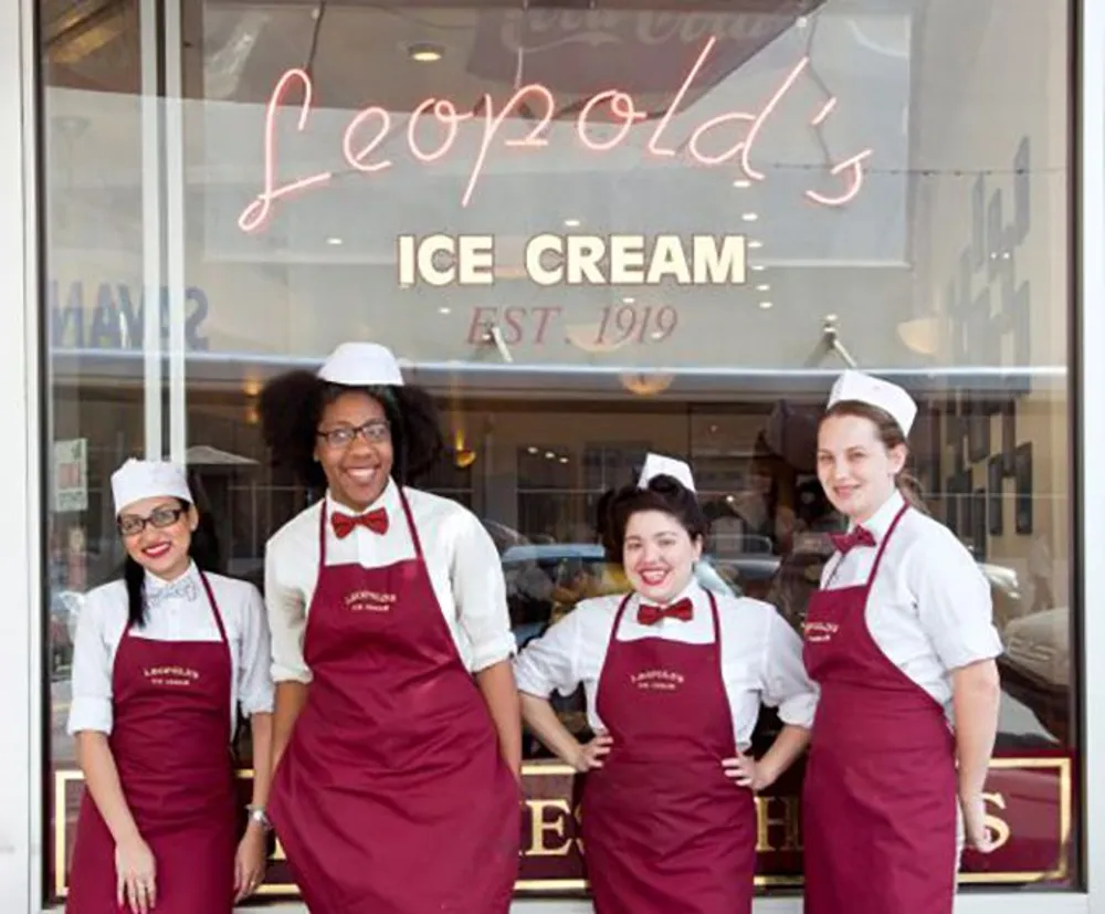 Four smiling employees in matching uniforms pose in front of Leopolds Ice Cream shop which boasts a vintage aesthetic with its cursive neon sign