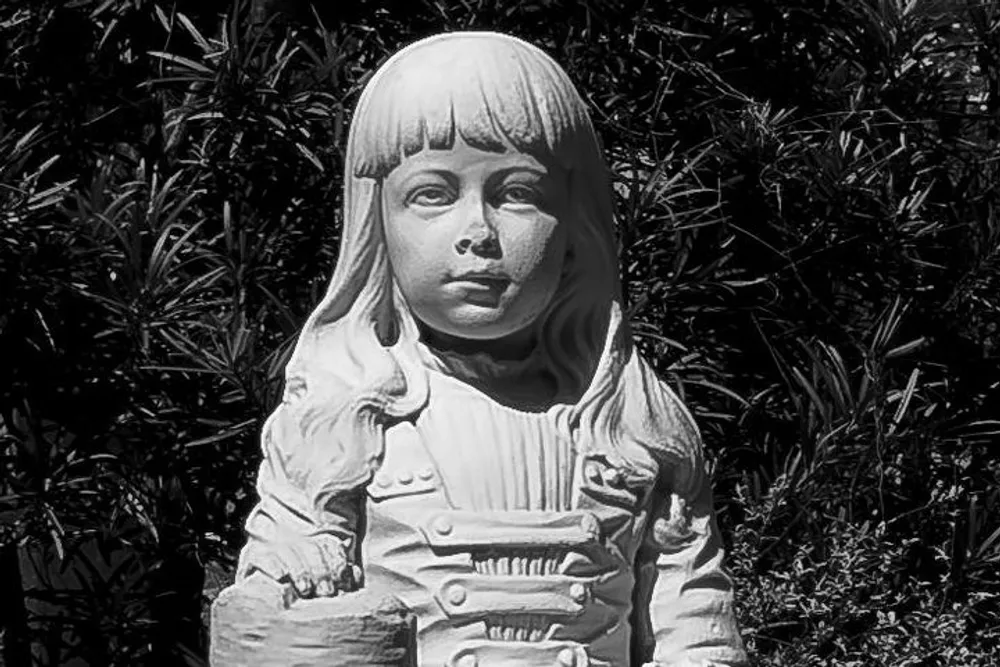 The image shows a black and white photo of a detailed sculpture depicting a young girl with a serene facial expression and mid-length hair set against a background of foliage