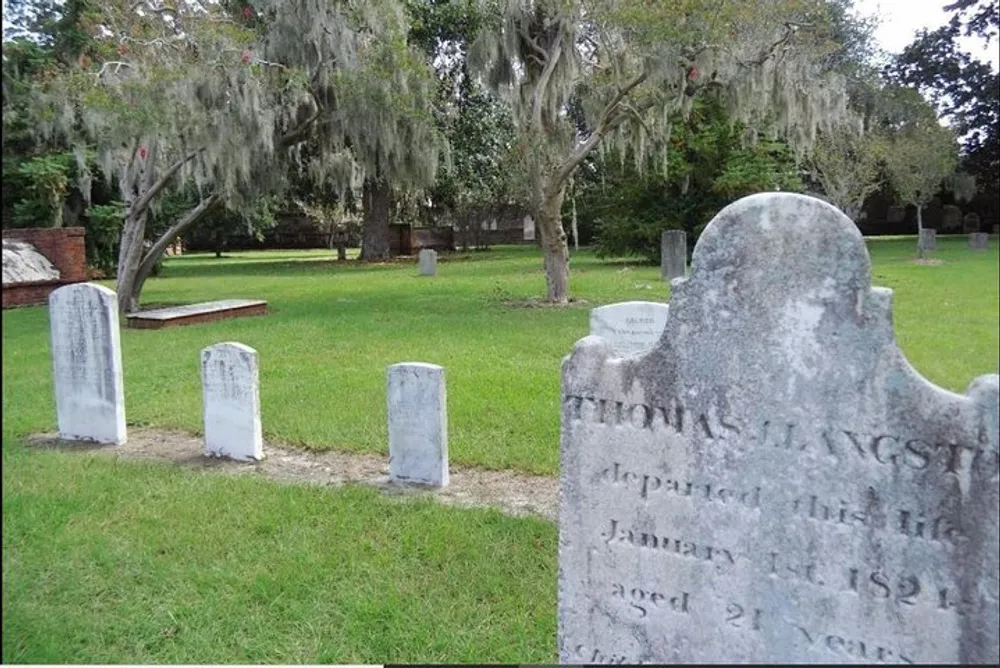 An old weathered gravestone is prominently displayed in a peaceful cemetery with multiple headstones and Spanish moss-draped trees in the background