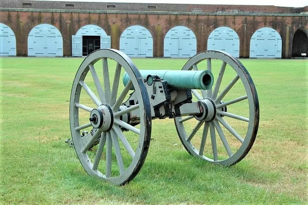 An old-fashioned cannon on wooden wheels is displayed on the grass in front of a building with rounded blue doors