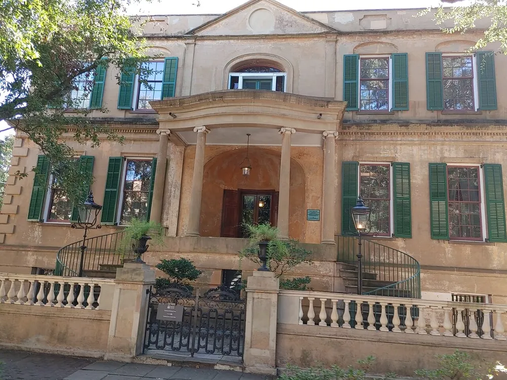 The image shows a two-story historic building with weathered stucco walls green shutters a curved entry staircase and an arched doorway exuding a classical architectural charm