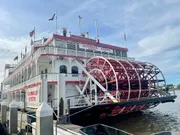 The image shows the Georgia Queen, a large red and white paddlewheel riverboat, docked at a pier under a blue sky.