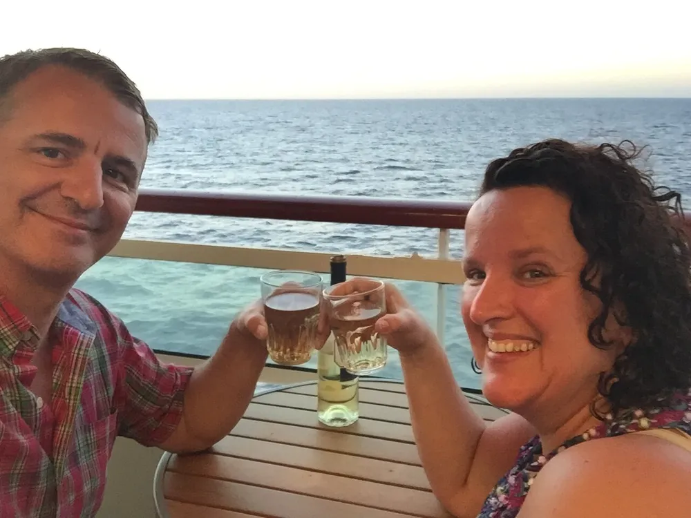 Two smiling individuals are toasting with glasses of wine on a balcony overlooking the sea at sunset