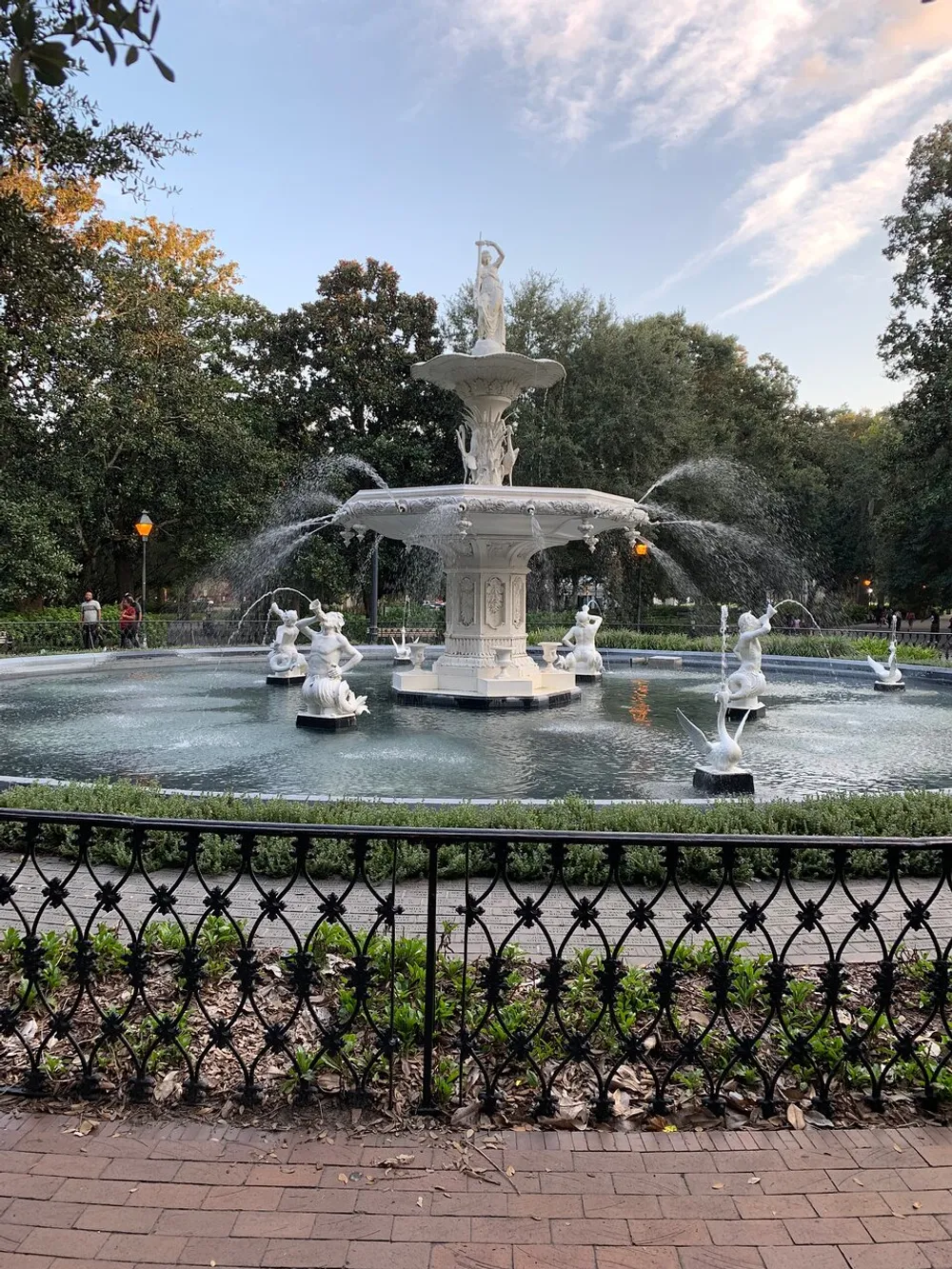 An ornate multi-tiered white fountain with water jets and statues is enclosed by a black wrought-iron fence in a park-like setting at dusk