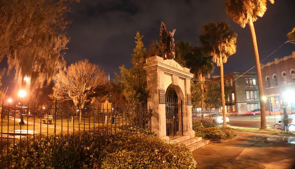 The image captures a night scene featuring an ornate gate with the acronym DAR at the entrance to a fenced area flanked by illuminated palm trees and moss-draped trees set against a backdrop of buildings and a dark sky