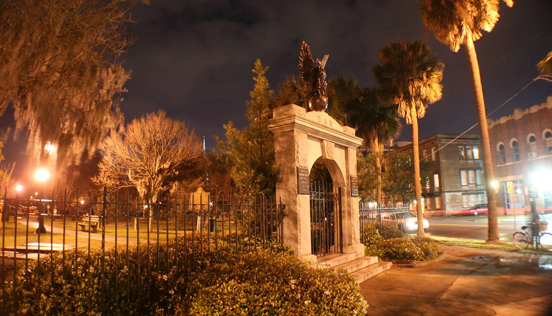 The image captures a night scene featuring an ornate gate with the acronym DAR at the entrance to a fenced area, flanked by illuminated palm trees and moss-draped trees, set against a backdrop of buildings and a dark sky.