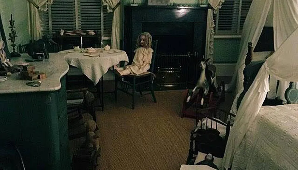 The image shows a dimly lit vintage room with a doll seated at a table creating a creepy or haunted atmosphere