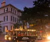 A Ghosts  Gravestones tour trolley is filled with passengers and travels through a street at dusk with a historic building to the left and trees draped with Spanish moss overhead
