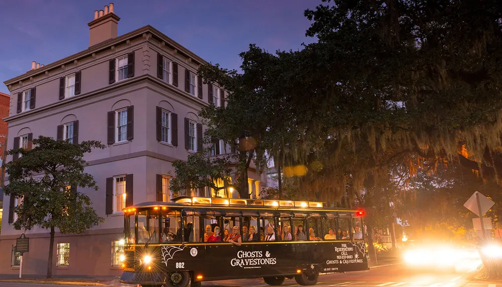 A Ghosts  Gravestones tour trolley is filled with passengers and travels through a street at dusk with a historic building to the left and trees draped with Spanish moss overhead