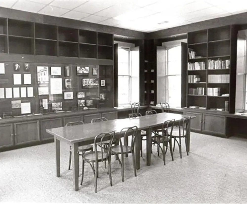 The image shows a vintage black and white photo of a quiet library or study room with a large table and chairs surrounded by bookshelves and bulletin boards
