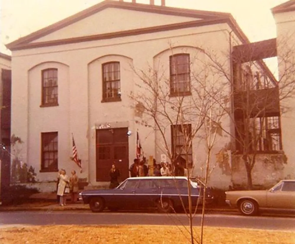The image shows a vintage photograph of several individuals gathered outside a historic building with two classic cars parked in front