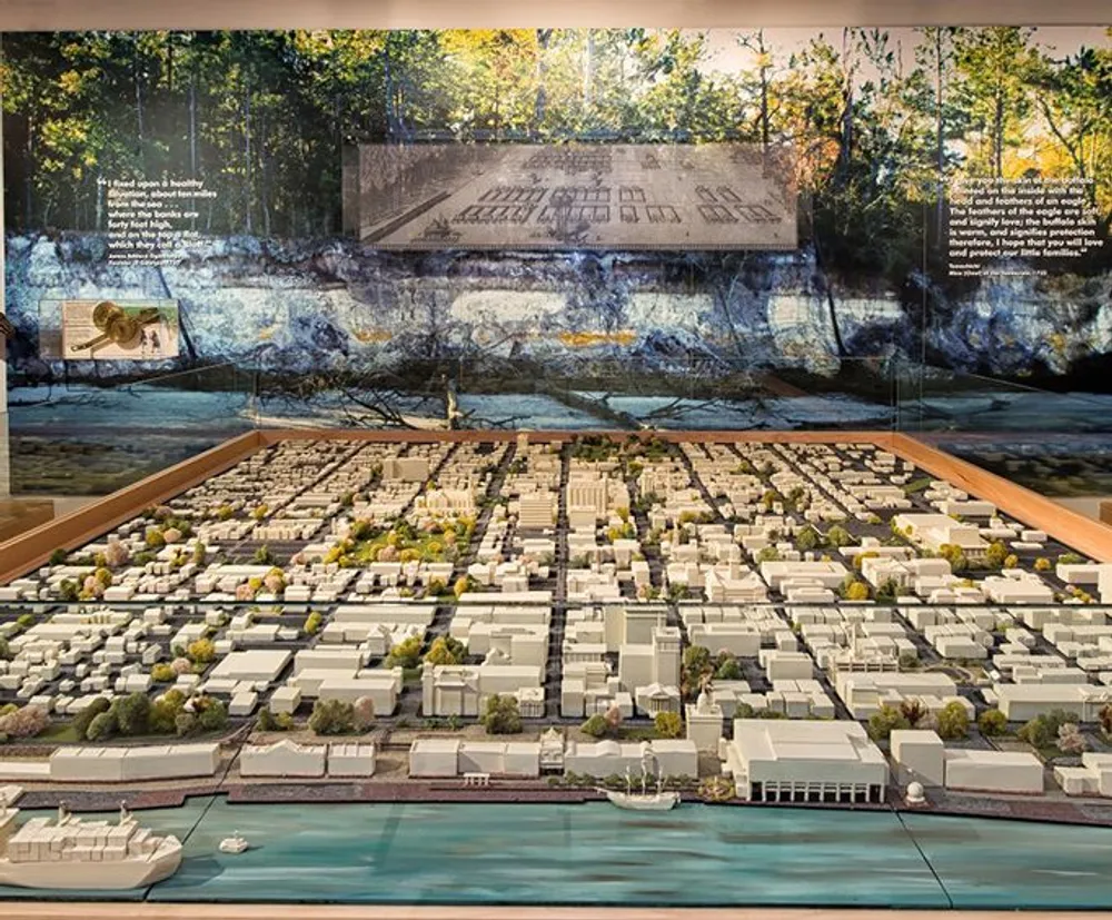 The image shows a detailed scale model of an urban area with buildings and streets set against a mural backdrop that blends images of nature and architectural plans