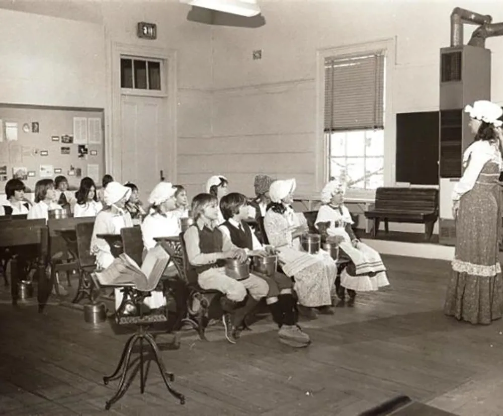 A group of children is dressed in historical costumes while seated inside what appears to be an old-fashioned classroom with a woman standing in period attire possibly teaching or conducting a lesson