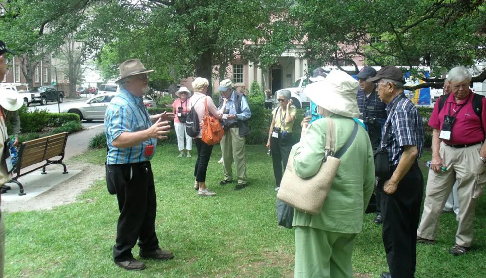 A tour guide wearing a hat is speaking to a group of attentive senior tourists in a park-like setting with greenery and benches