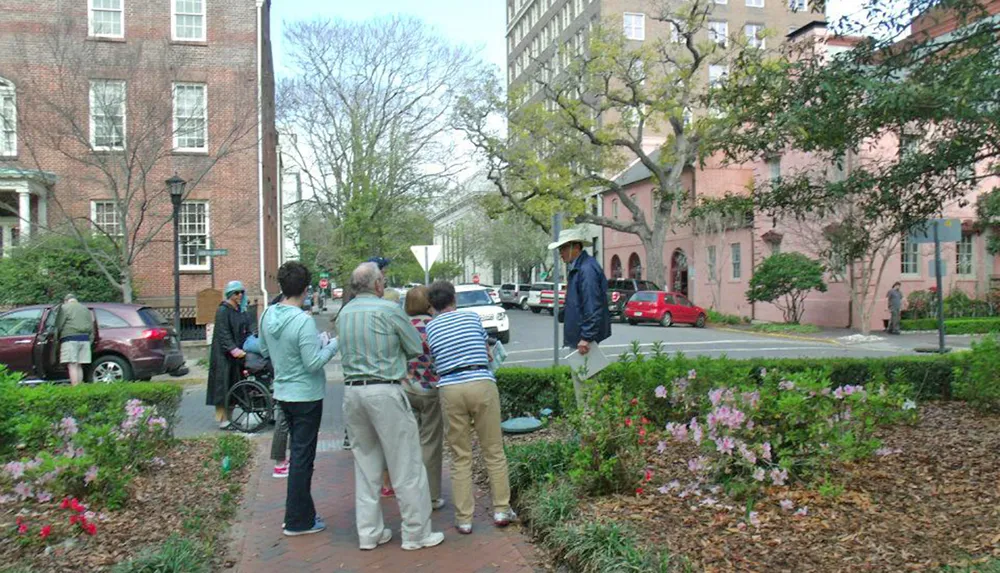 A group of people one in a wheelchair appear to be listening to a guide wearing a hat possibly on an outdoor tour in an urban setting with flowering bushes in the foreground