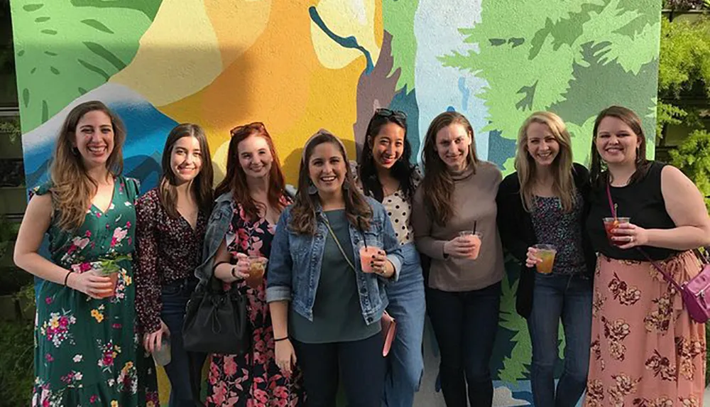 A group of smiling women pose for a photo in front of a colorful mural with beverages in their hands