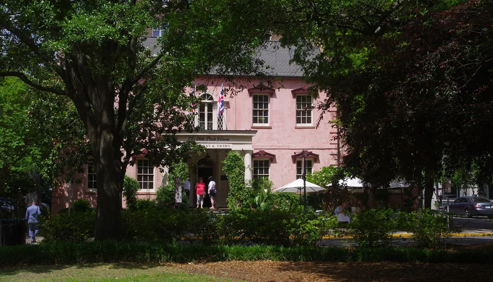The photo features a charming pink historical building surrounded by lush trees under a clear blue sky with people visible near the entrance
