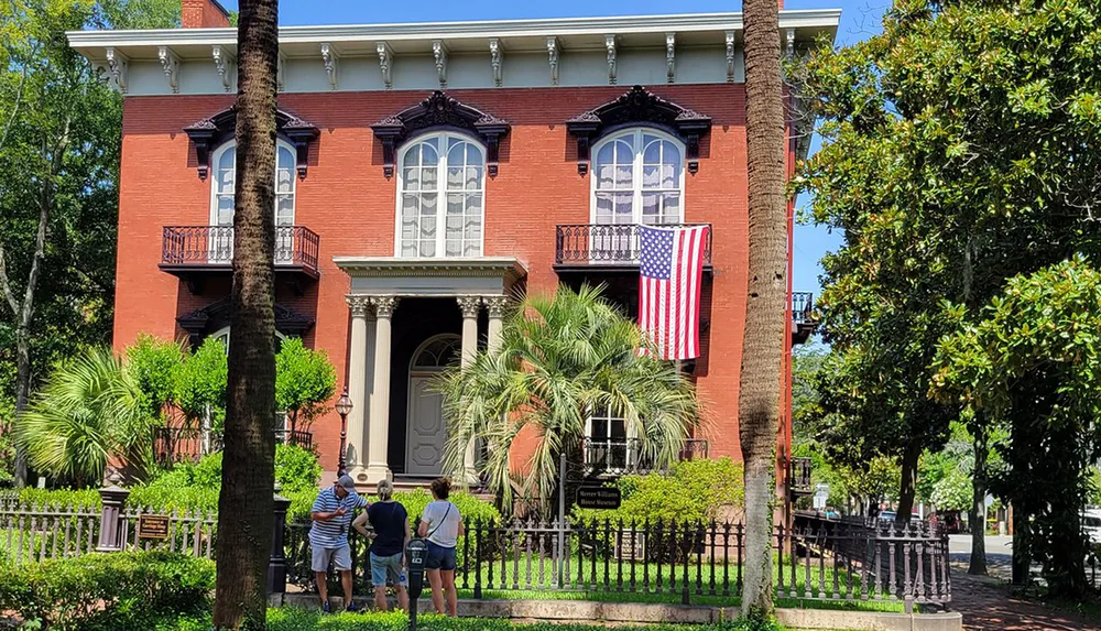 A group of people is standing outside a stately red brick house adorned with an American flag on a sunny day surrounded by greenery