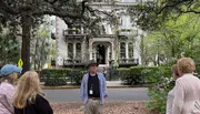 A group of people are standing in a park with Spanish moss-laden trees in front of an elegant building with balconies and flowering plants.