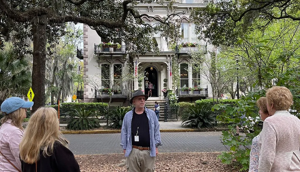 A group of people are standing in a park with Spanish moss-laden trees in front of an elegant building with balconies and flowering plants
