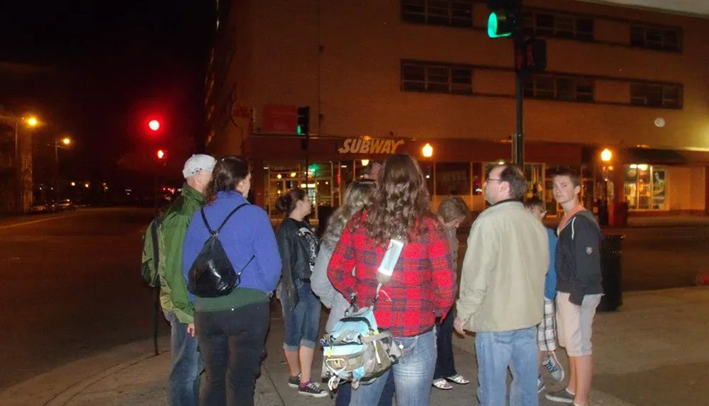 A group of people is standing at a street corner during the evening with a traffic light showing red and a Subway restaurant sign visible in the background