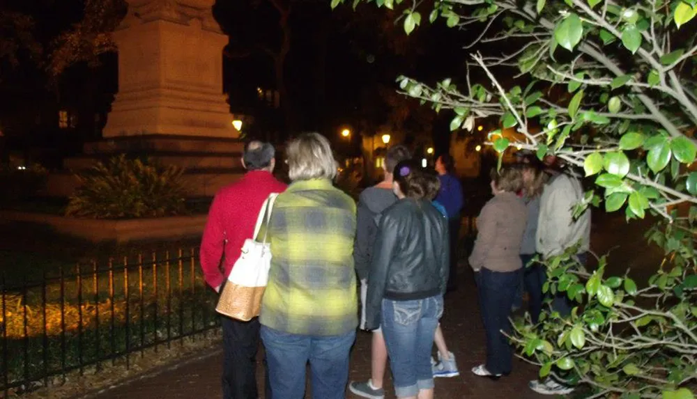 A group of people is standing outdoors at night possibly listening to a speaker or looking at something of interest in a park-like setting with trees and a monument