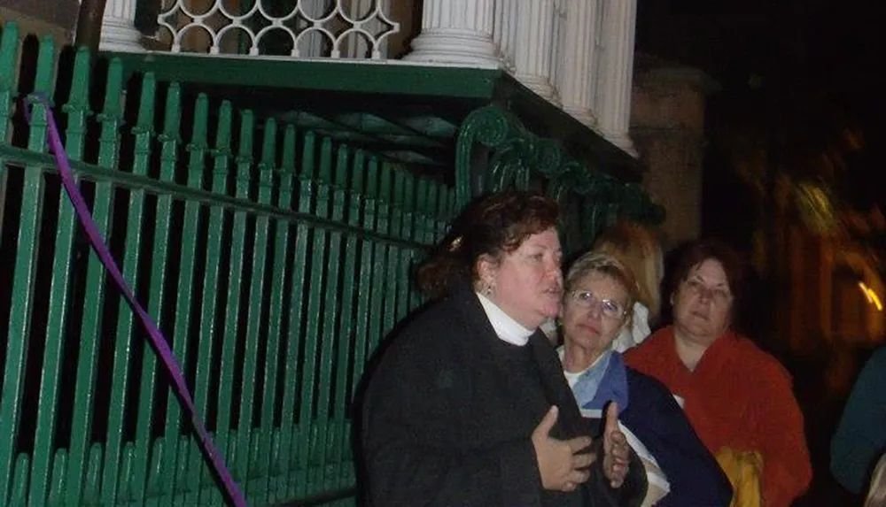 The image shows a group of people with a woman in the foreground seemingly speaking or gesturing standing near a green fence at night
