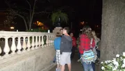 The image shows a group of people, possibly on a tour, gathered at night near a stone balustrade, with a focus on a young person wearing a grey hoodie looking across the railing.