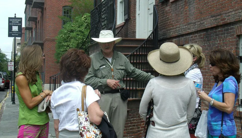 A tour guide in a wide-brimmed hat is explaining something to a group of attentive women on a city street