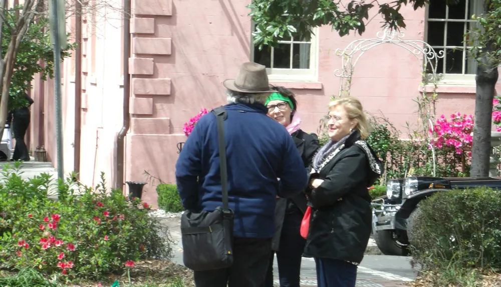 Three individuals are engaged in a conversation in front of a pink building adorned with vibrant flowers