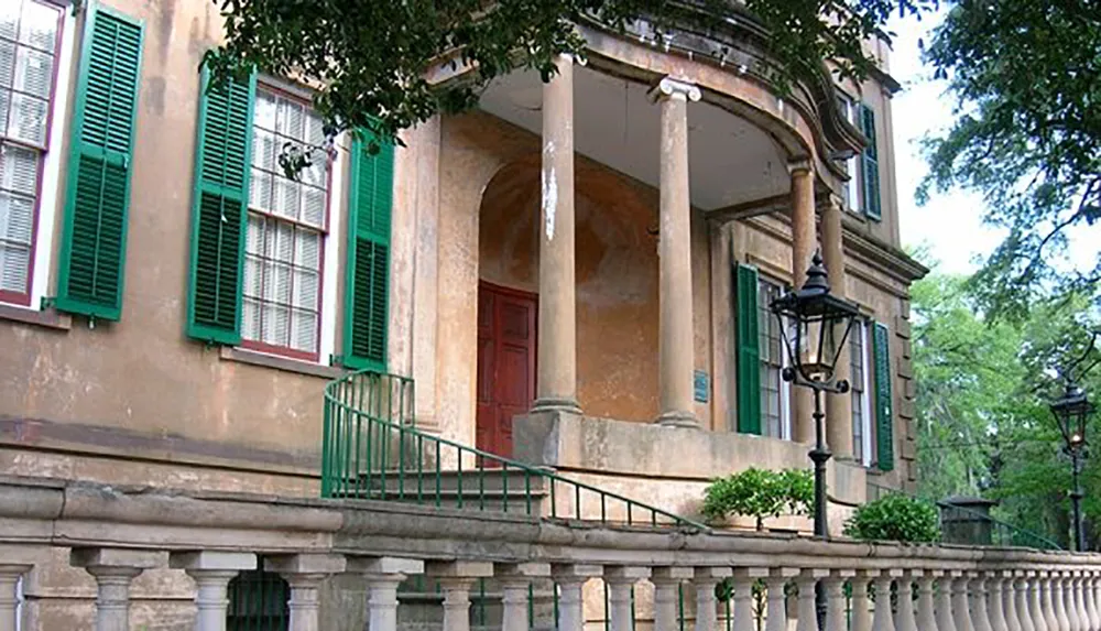 The image shows an old building with green window shutters a red door and a curved portico featuring classic architectural details like columns and balustrades accompanied by vintage-style lanterns